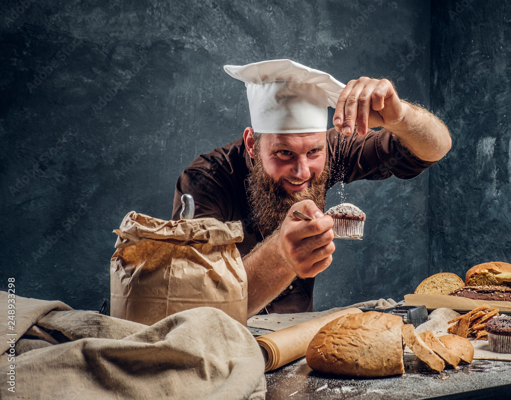 Cheerful bearded baker sprinkling some flour on his freshly made muffin next to a table in a dark studio