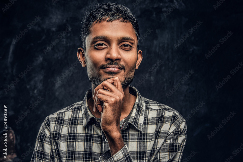 Fototapeta premium Close-up portrait of a handsome young Indian guy wearing a checkered shirt holding hand on chin and looking at a camera with a pensive look.