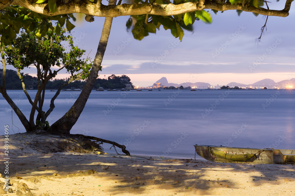 Rio de Janeiro City view and Guanabara Bay with Sugar Loaf in the background from Paqueta Island, Brazil. Fisherman's boat and tree in the foreground. Shades of lavender at sunset. 2017 Rio de Janeiro City view and Guanabara Bay with Sugar Loaf in the background from Paqueta Island, Brazil. Fisherman's boat and tree in the foreground. Shades of lavender at sunset. 2017