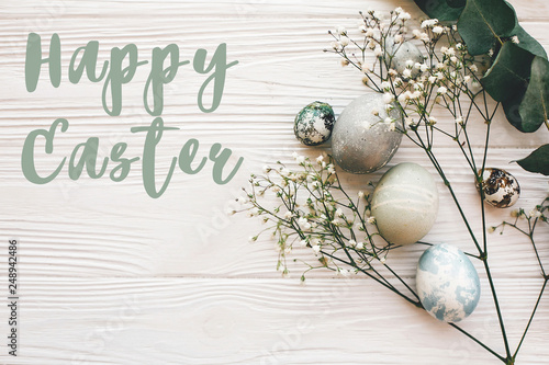 Happy Easter text sign on stylish easter eggs with spring flowers and green branch on white wooden table, flat lay. Modern easter eggs painted with natural dye. Easter greetings card