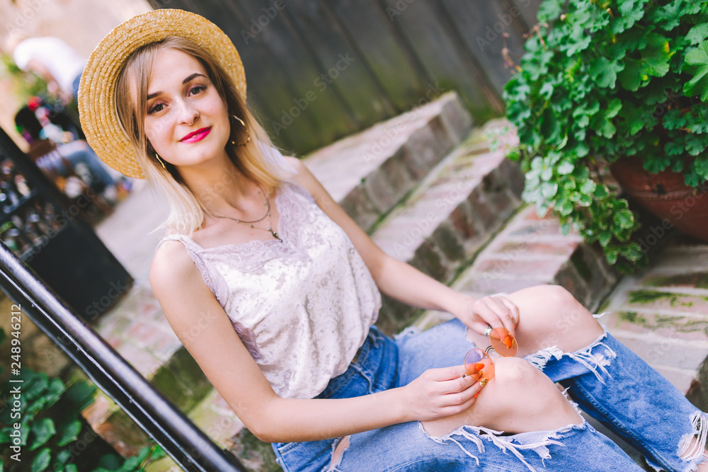 Attractive blonde girl with beautiful smile. A beautiful woman dressed in a straw hat and blue jeans sitting on the stairs.