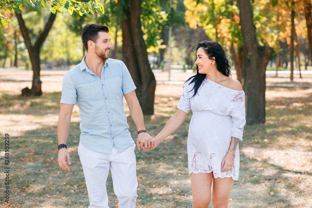 Pregnant girl walks in the park with her husband, hugging and kissing, enjoying the beautiful weather Pregnant girl walks in the park with her husband, hugging and kissing, enjoying the beautiful weather
