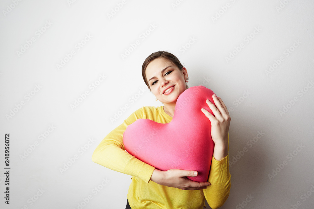 Young beautiful girl in yellow tshirt holding big plush red heart over empty white background.Lifestyle and Holiday Concept Young beautiful girl in yellow tshirt holding big plush red heart over empty white background.Lifestyle and Holiday Concept