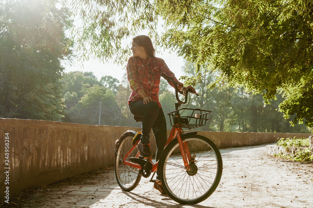 Subject ecological bicycle transport. Young caucasian woman on a dirt road in a park near a lake renting an orange-colored bike posing for a rest in the fall in sunny weather