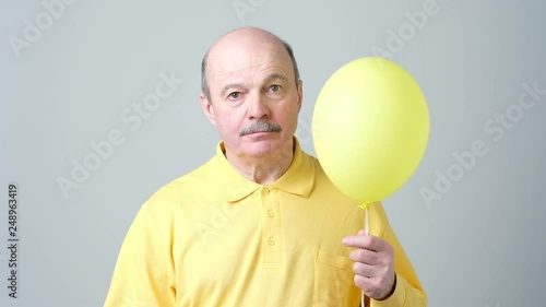 A sad senior man in a yellow shirt is holding a small balloon in his hands.