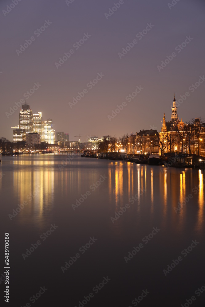 AMSTERDAM, NETHERLANDS - NOVEMBER 21, 2018: Illuminated buildings reflected in calm water, amazing cityscape with boat on Amstel canal at night
