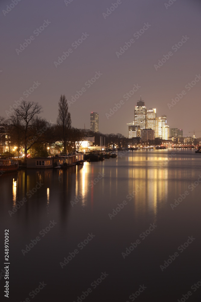 AMSTERDAM, NETHERLANDS - NOVEMBER 21, 2018: Illuminated buildings reflected in calm water, amazing cityscape with boat on Amstel canal at night