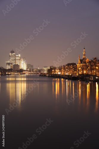 AMSTERDAM, NETHERLANDS - NOVEMBER 21, 2018: Illuminated buildings reflected in calm water, amazing cityscape with boat on Amstel canal at night