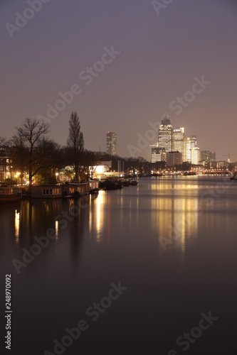 AMSTERDAM  NETHERLANDS - NOVEMBER 21  2018  Illuminated buildings reflected in calm water  amazing cityscape with boat on Amstel canal at night