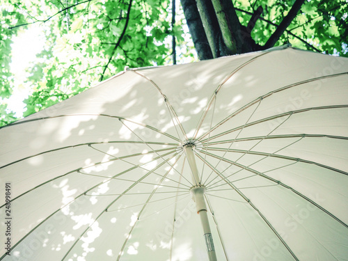 White sun umbrella in a summer day light. sunshine, with green forest park background