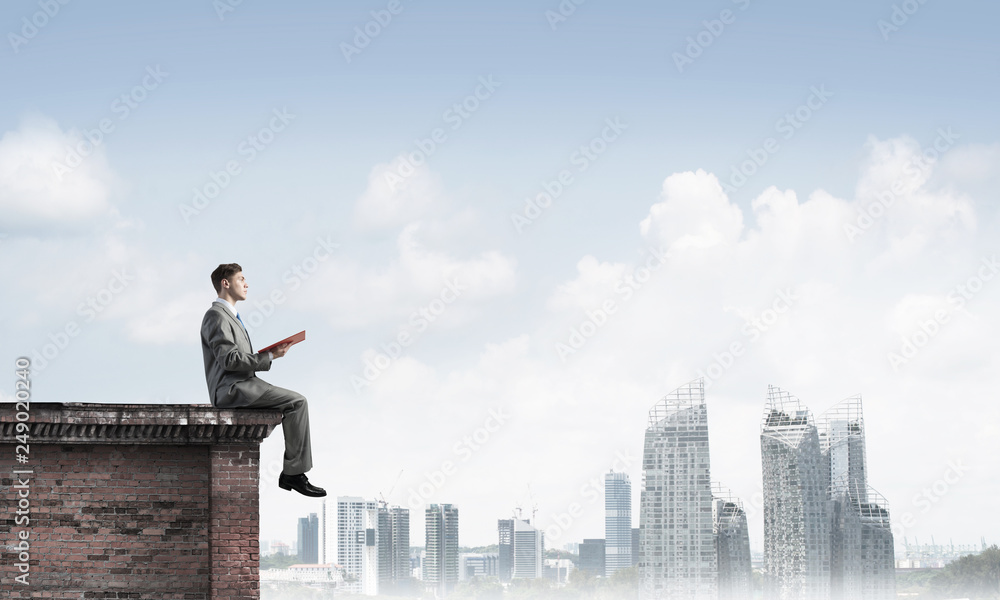 Man on roof edge reading book and cityscape at background Man on roof edge reading book and cityscape at background
