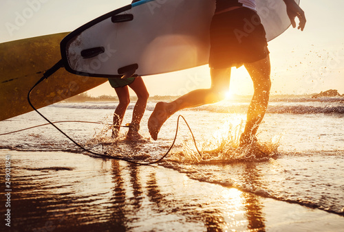 Son and father surfers run in ocean waves with long boards. Close up splashes and legs image