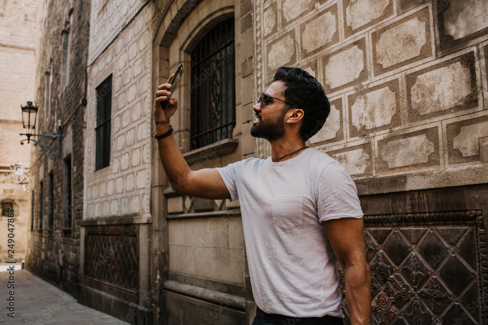 Content attractive hispanic man in white tshirt photographing himself on mobile phone while enjoying city travel.Bearded hipster male making selfie via smartphone on gotic streets