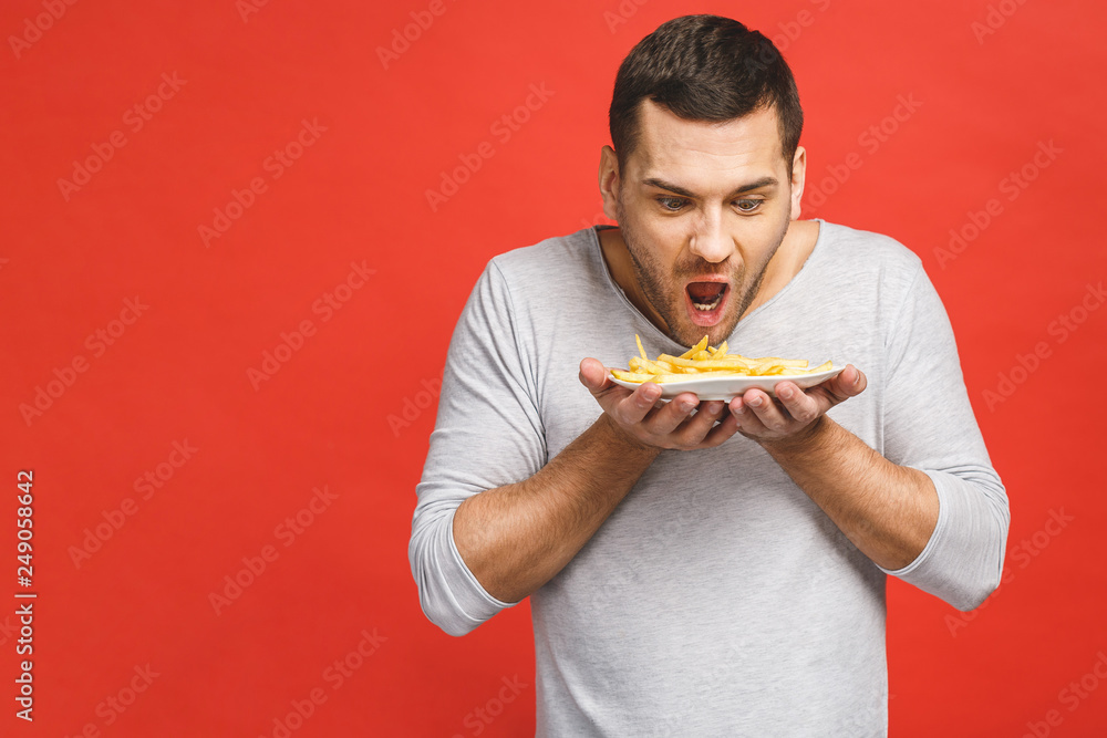 Young man holding french fries. Student eats fast food. Not helpful food. Very hungry guy. Diet concept. Isolated over red background. Young man holding french fries. Student eats fast food. Not helpful food. Very hungry guy. Diet concept. Isolated over red background.