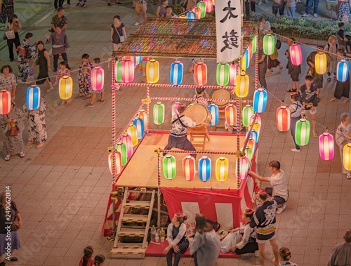 View of the square in front of the Nippori train station decorated for the Obon festival with a yagura tower illuminated with paper lanterns where a girl in traditional costume is playing taiko drum.