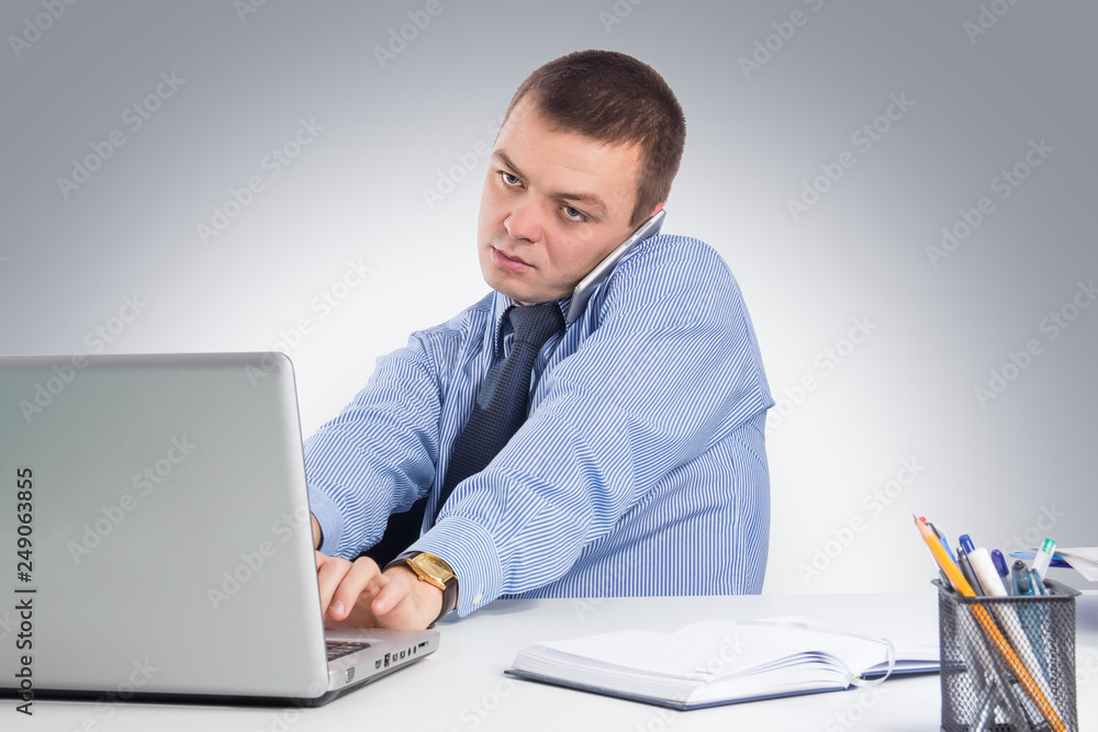 Businessman with smartphone typing on laptop in his office on gray background Businessman with smartphone typing on laptop in his office on gray background