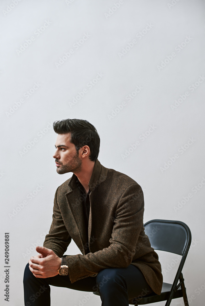 Confident and handsome. Fashionable young man sitting on a chair with his hands on his knees, looking away isolated over white background Confident and handsome. Fashionable young man sitting on a chair with his hands on his knees, looking away isolated over white background