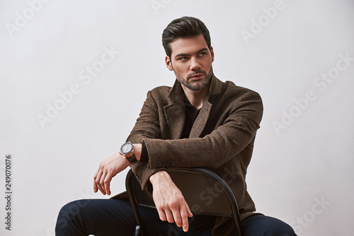 Perfect style. Stylish dark-haired man sitting on a chair and looking away isolated over white studio background