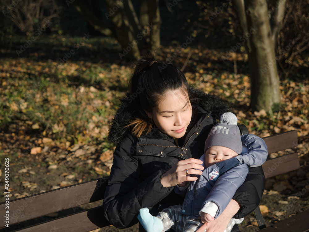 Fototapeta premium Happy Asian young mother walking with baby outdoors in the park during Autumn, good cold weather, happy child, mixed race family, half Asian Half European. Autumn scene of mother kissing young baby