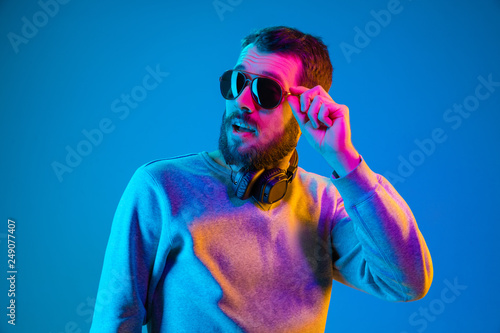Enjoying his favorite music. Happy young stylish man in hat and sunglasses with headphones listening and smiling while standing against blue neon background photo
