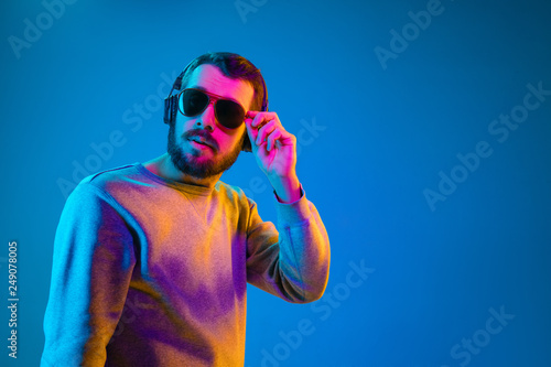 Enjoying his favorite music. Serious young stylish man in sunglasses with headphones listening sound while standing against blue neon background
