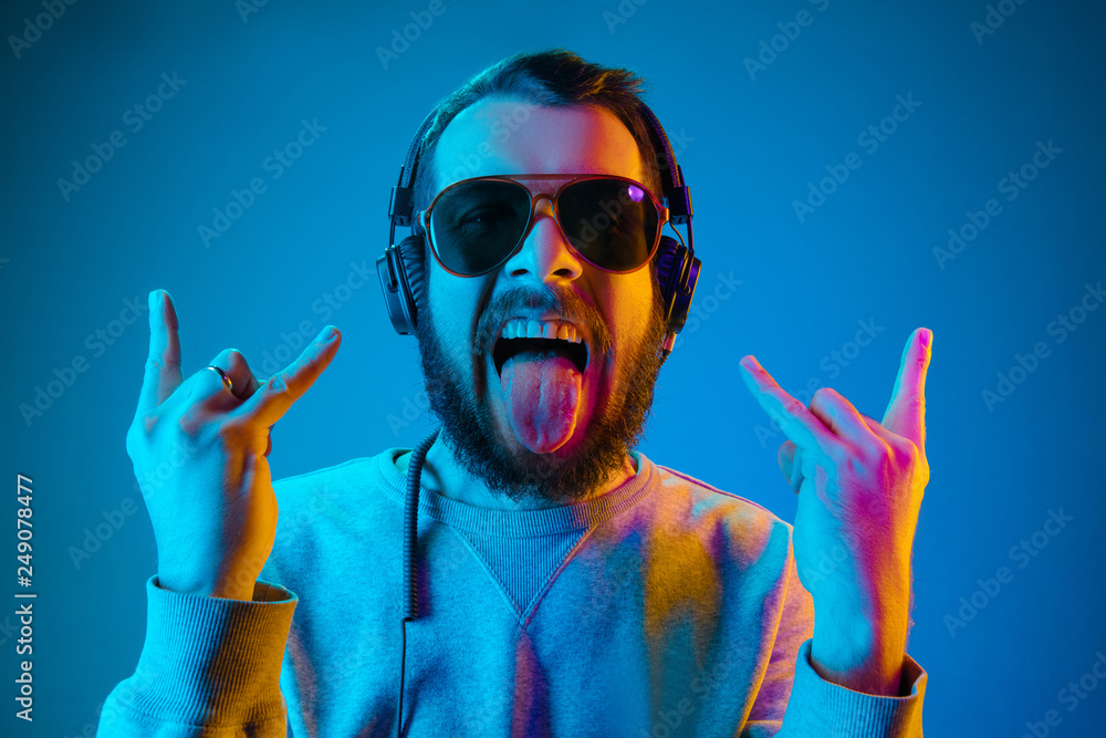 Enjoying his favorite music. Happy young stylish man in sunglasses with headphones listening sound and smiling while standing against blue neon background - obrazy, fototapety, plakaty 