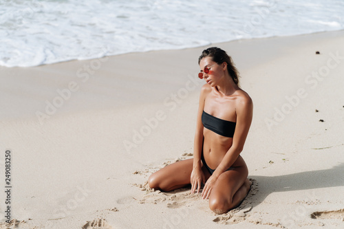 Sexy tanned girl in black swimsuit and sunglasses posing on sandy beach