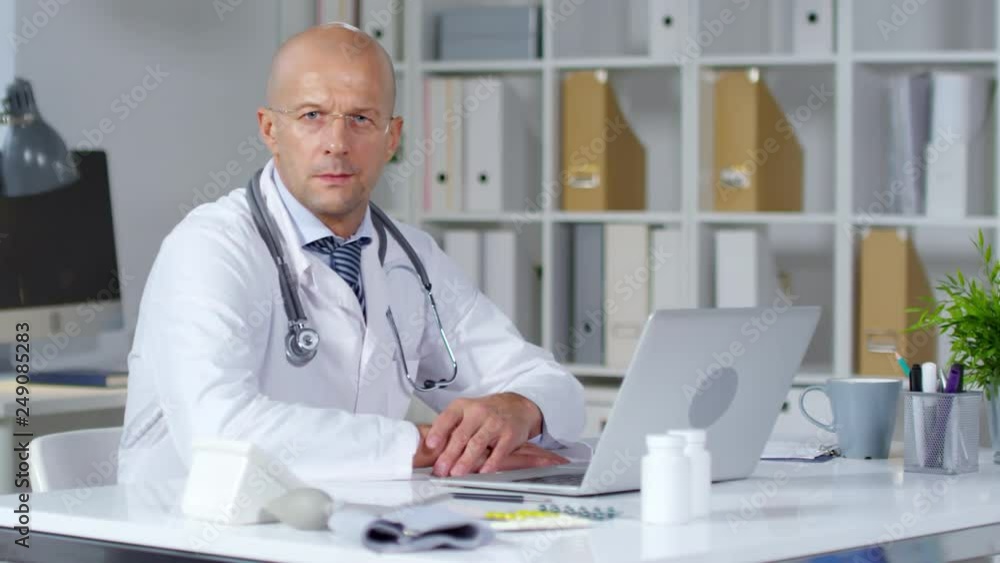 Medium shot of bald male doctor in white coat and glasses sitting at his desk and working laptop, then turning towards camera and posing Medium shot of bald male doctor in white coat and glasses sitting at his desk and working laptop, then turning towards camera and posing