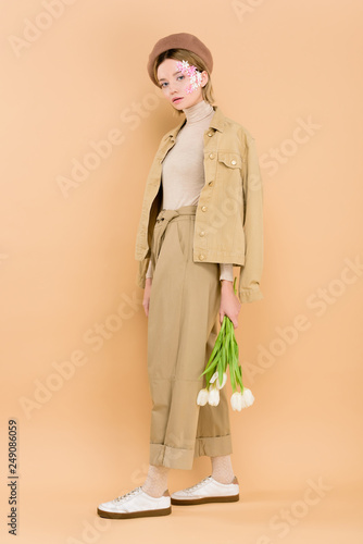 trendy woman in beret holding bouquet isolated on beige