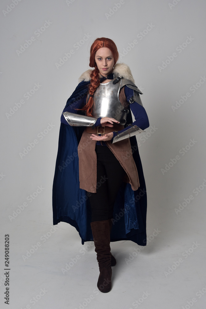 Naklejka premium full length portrait of a red haired girl wearing medieval warrior costume and steel armour and a fur cloak, standing pose on grey studio background.