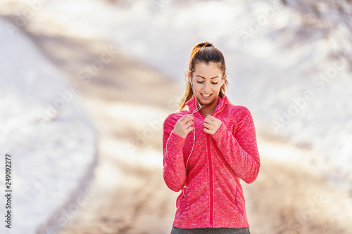 Quadro em tela Smiling sporty woman zipping sweatshirt while standing on the trail at winter