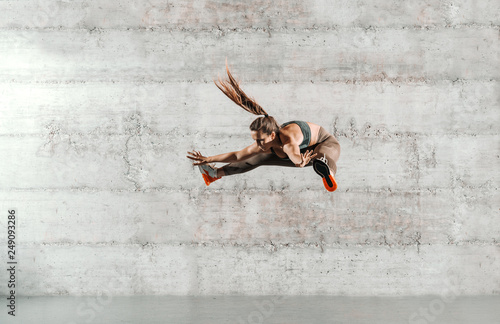 Caucasian woman in sportswear and with ponytail jumping with spread arms and legs. In background wall. Full length.