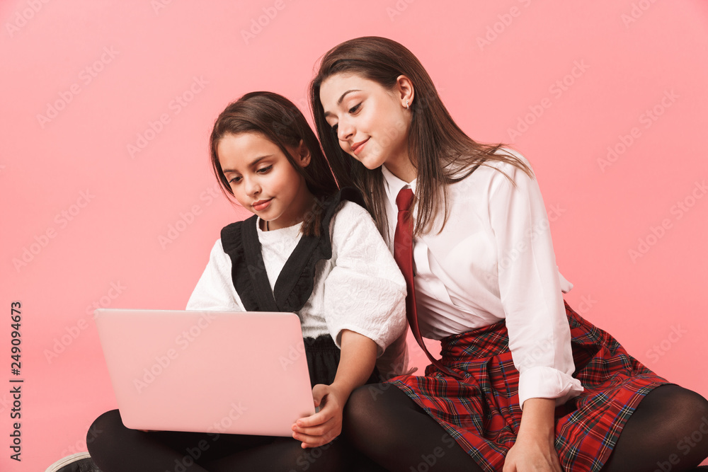 Portrait of little girls in school uniform using laptop, while sitting on floor isolated over red background Portrait of little girls in school uniform using laptop, while sitting on floor isolated over red background