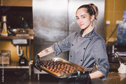 Subject profession and cooking pastry. young Caucasian woman with tattoo of pastry chef in kitchen of restaurant preparing round chocolate candies handmade truffle in black gloves and uniform