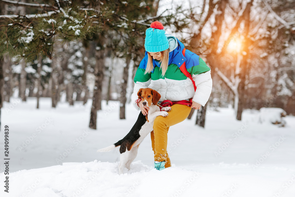 Portrait of cheerful young woman in warm stylish clothes and bagpack posing with her beagle dog in winter park. Friendship, pet and human.