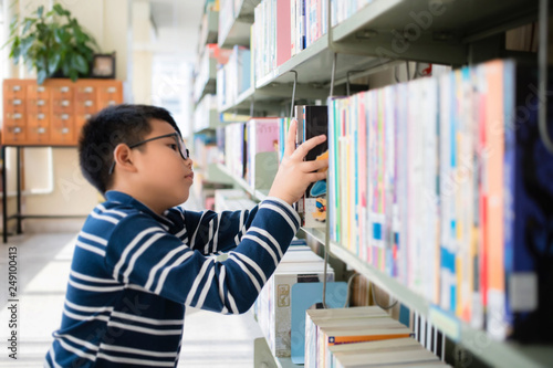Asian Thai boy wearing glasses, long sleeves and shorts. He is choosing and picking up books on the bookshelf. in the library at school. In Bangkok, Thailand.