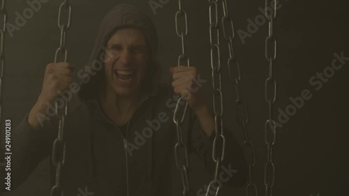 Tough man in hoodie trying hard to break rusty iron chains to liberate himself from shackles of dogma and social prejudices over black background with smoke. Guy tearing chains to free his soul .