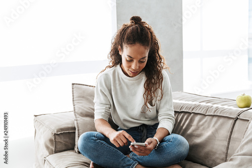 Photo of adorable african american woman using cell phone, while sitting on couch in bright apartment