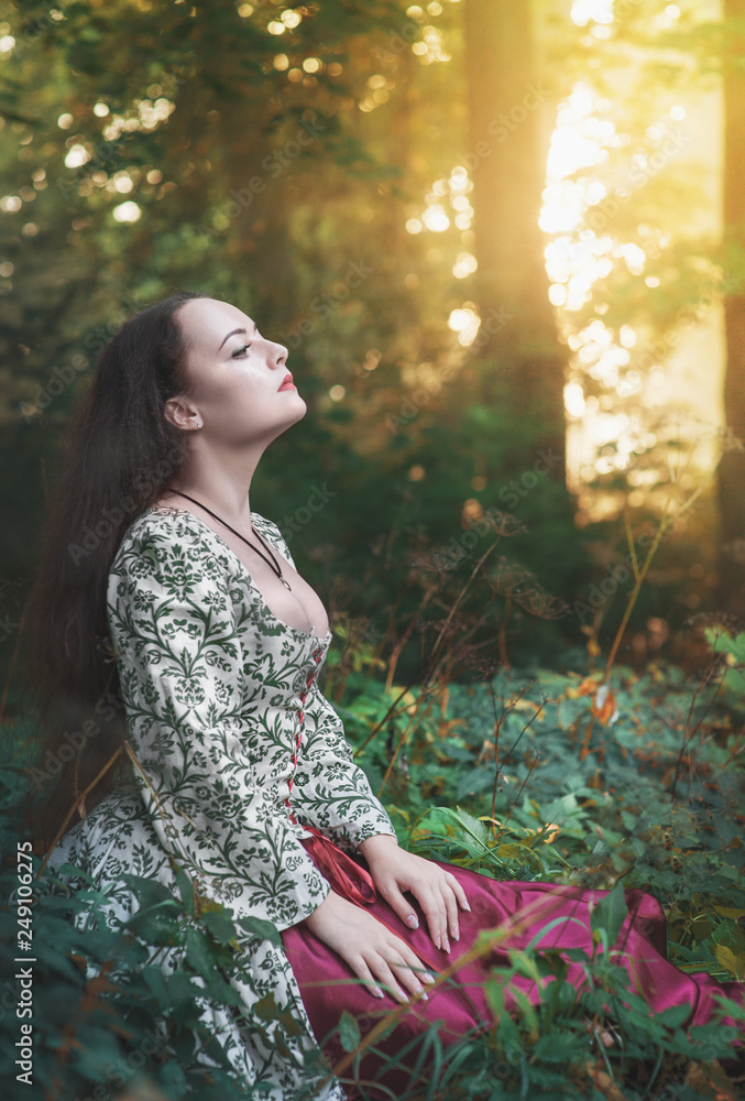 Beautiful woman in medieval dress sitting in grass with sunset lights Beautiful woman in medieval dress sitting in grass with sunset lights