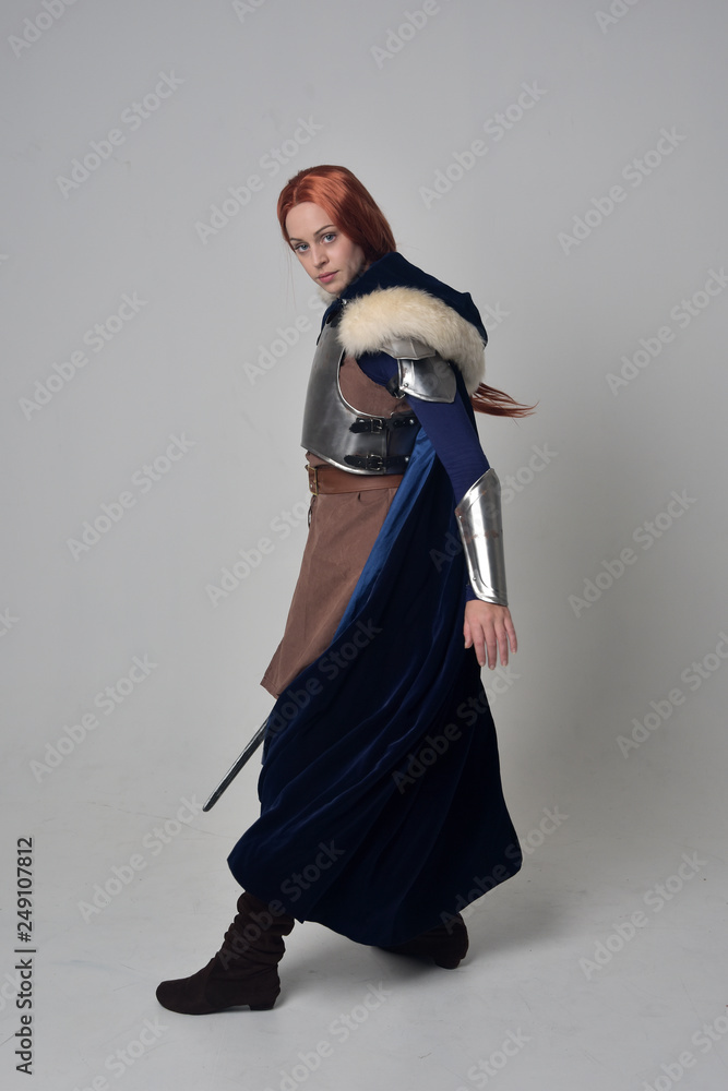 full length portrait of a  red haired girl wearing medieval warrior costume and steel armour and a fur cloak, standing pose on grey studio background.