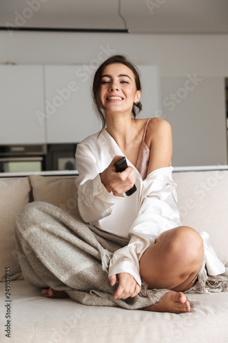 Portrait of joyful woman 20s in leisure wear, holding remote control while sitting on couch in bright flat