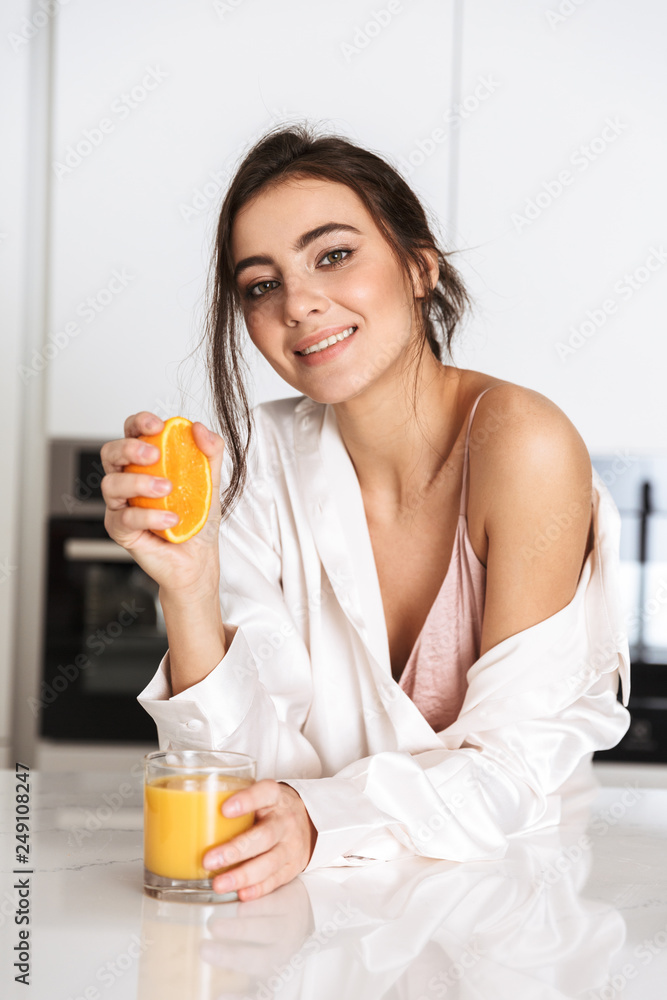Image of european woman 20s wearing silk leisure clothing, making orange juice with fruit in kitchen Image of european woman 20s wearing silk leisure clothing, making orange juice with fruit in kitchen