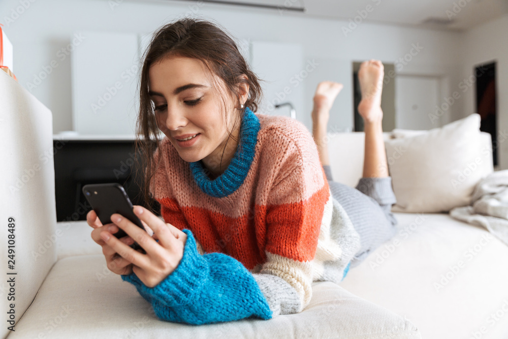 Photo of beautiful woman wearing sweater, lying on sofa in apartment and using cell phone Photo of beautiful woman wearing sweater, lying on sofa in apartment and using cell phone