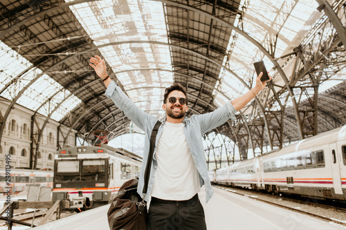 Attractive positive businessman feeling himself happy after long business travel trip.Casual professional entrepreneur using smartphone at hall of train station