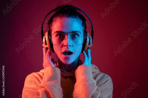 Surprised pretty woman with headphones listening to music over red neon background at studio.