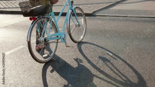 A woman walks a vintage bicycle across a road with long shadows at golden hour