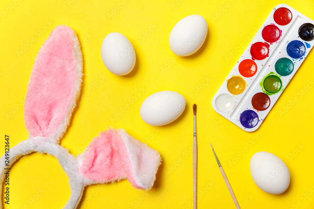 Happy Easter concept. Preparation for holiday. Decorative eggs colorful paints and bunny ears furry costume toy isolated on trendy yellow background. Simple minimalism flat lay top view copy space Happy Easter concept. Preparation for holiday. Decorative eggs colorful paints and bunny ears furry costume toy isolated on trendy yellow background. Simple minimalism flat lay top view copy space