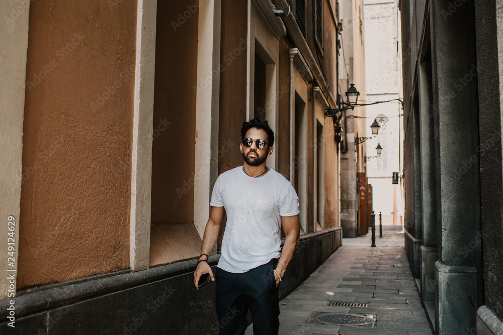 Content attractive hispanic man in white tshirt walking old town streets holding mobile phone hand.Bearded hipster male enjoying city travel on gotic district streets Content attractive hispanic man in white tshirt walking old town streets holding mobile phone hand.Bearded hipster male enjoying city travel on gotic district streets