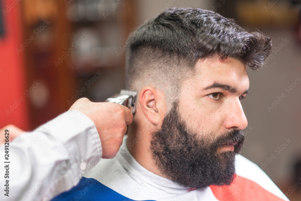 Fototapeta premium Handsome bearded man, getting haircut by barber, with electric trimmer at barbershop .