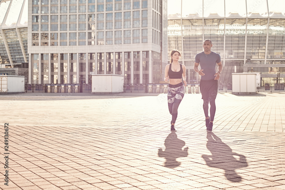Obraz premium Positive and sporty mixed race couple: handsome african man and brunette girl in sportswear running together on a sunny warm day.